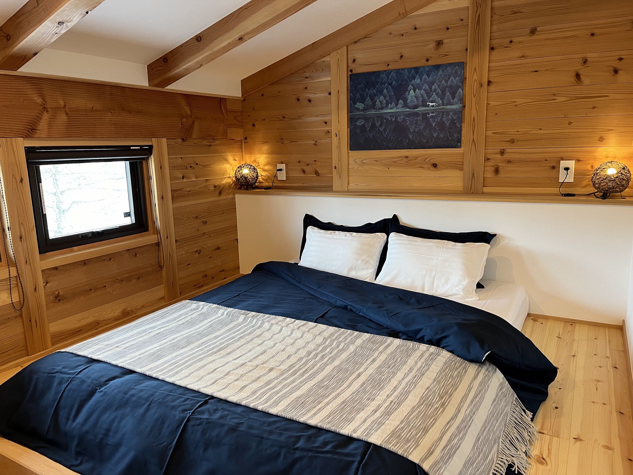 Main bedroom with a double bed in a navy duvet, warm bedside lamps and a forest painting on the timber wall