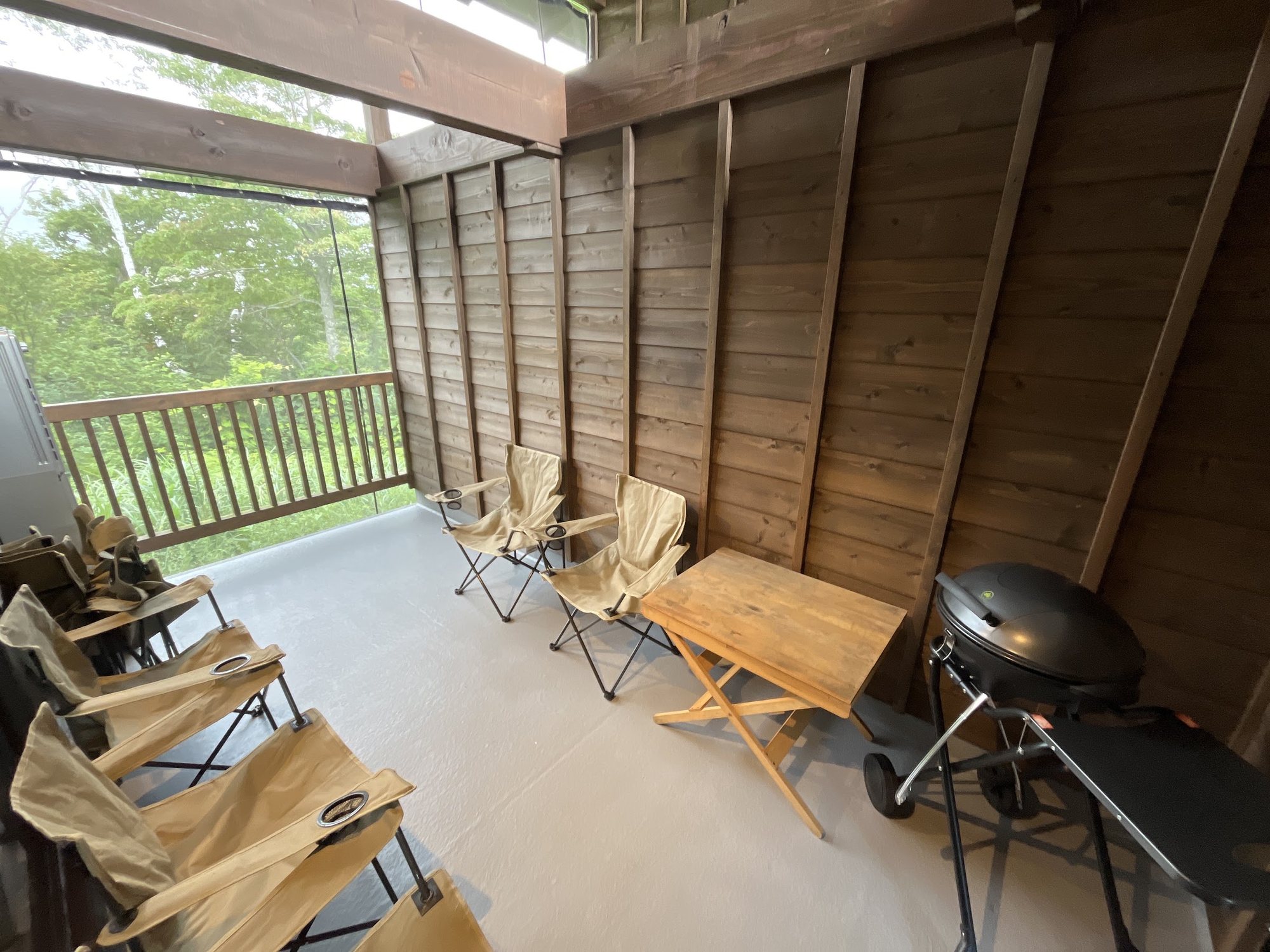 Covered deck with camping chairs, a folding table and a BBQ grill, green forest beyond