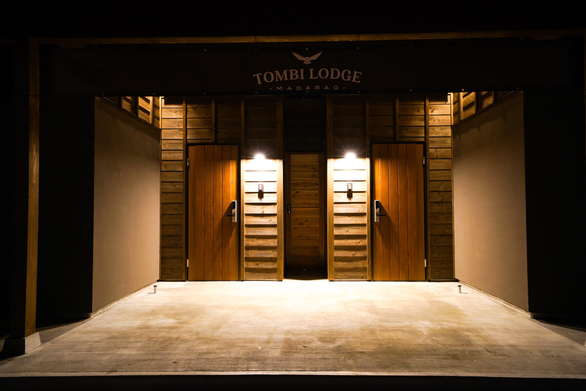 The Tombi Lodge entrance at night, its two wooden front doors lit by wall lamps under the Tombi Lodge Madarao sign
